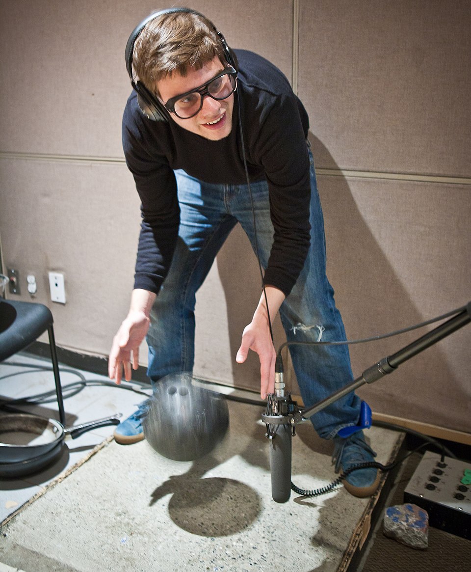 A Foley artist wearing headphones drops a bowling ball near a microphone in a sound studio