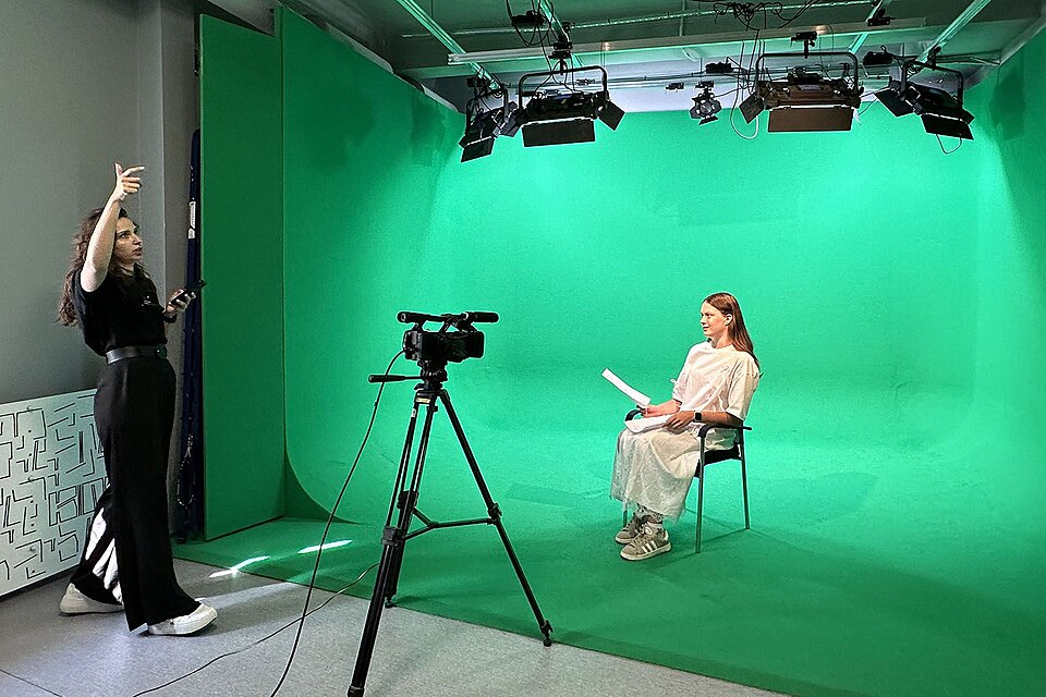 A film studio with a bright green screen backdrop, camera on a tripod, and studio lights overhead