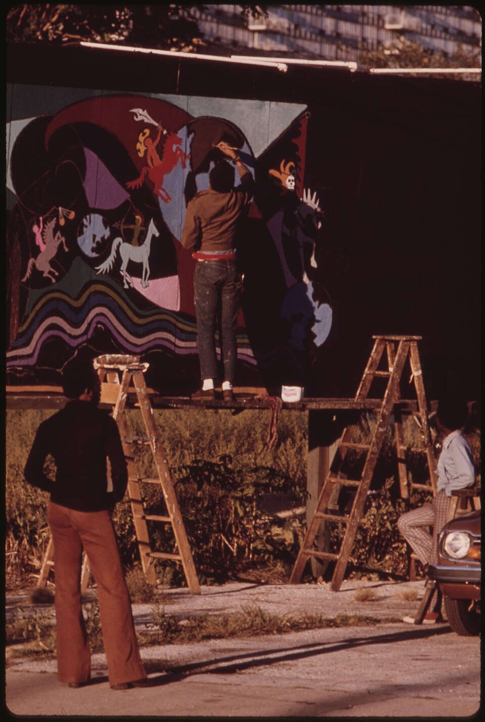 An artist standing on a scaffold between two ladders, painting a colorful mural on a wall