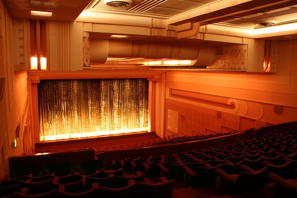 The inside of a movie theater with rows of red seats and a big curtained screen