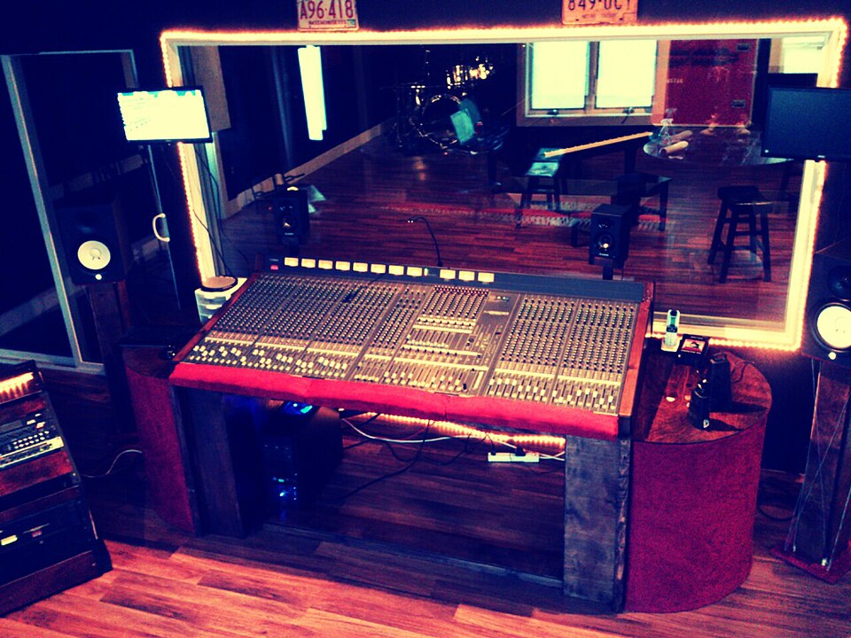 A recording studio control room with a mixing console and a window looking into the recording space