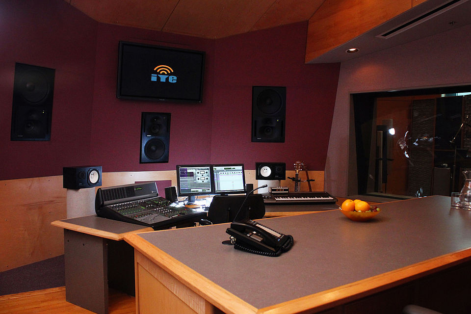 A sound mixing studio with a mixing console, speakers, and a window into the recording room