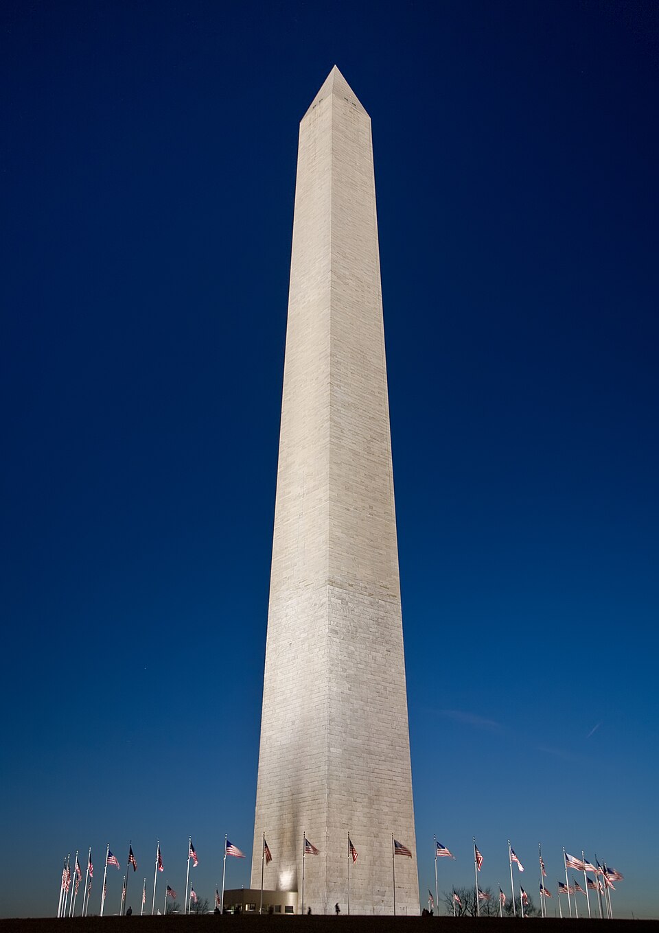 The Washington Monument rising tall against a deep blue sky at dusk, surrounded by American flags